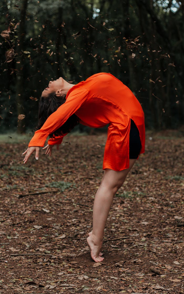 Woman in vibrant orange performing a dynamic yoga pose set in a tranquil forest scene.